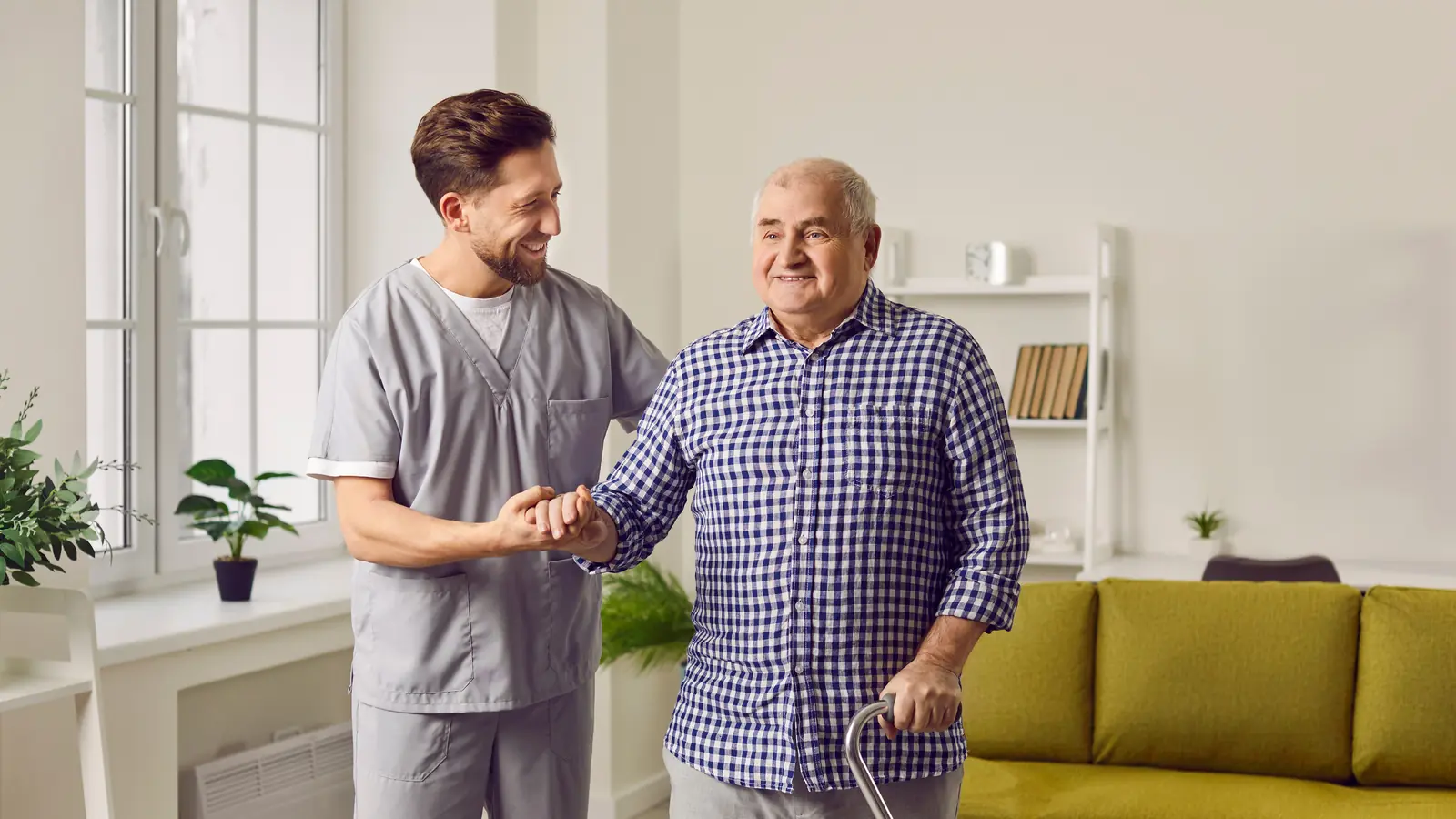 Caregiver helping a smiling senior man with a cane walk through his living room