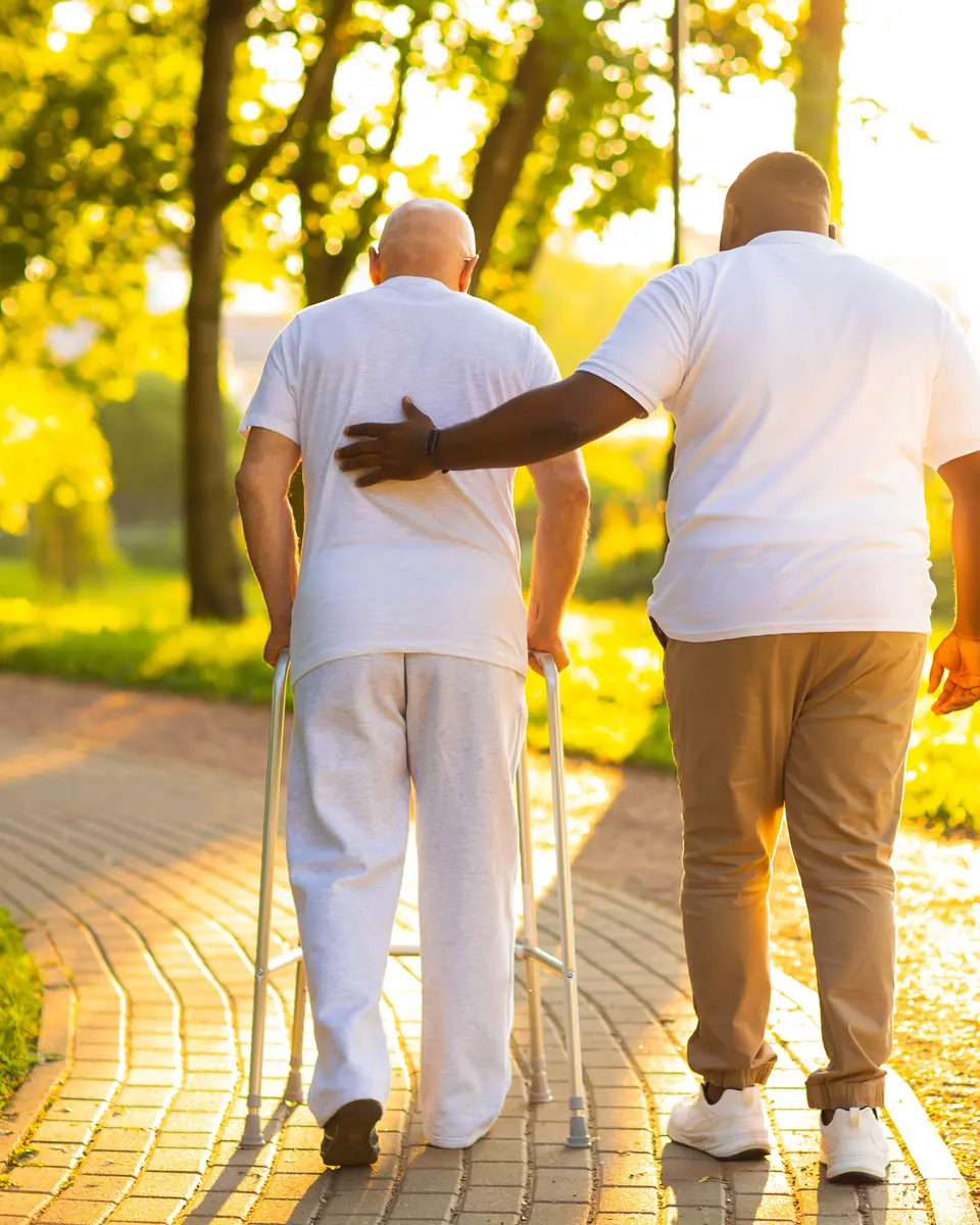 Caregiver supporting a senior man with a walker on a sunny park path