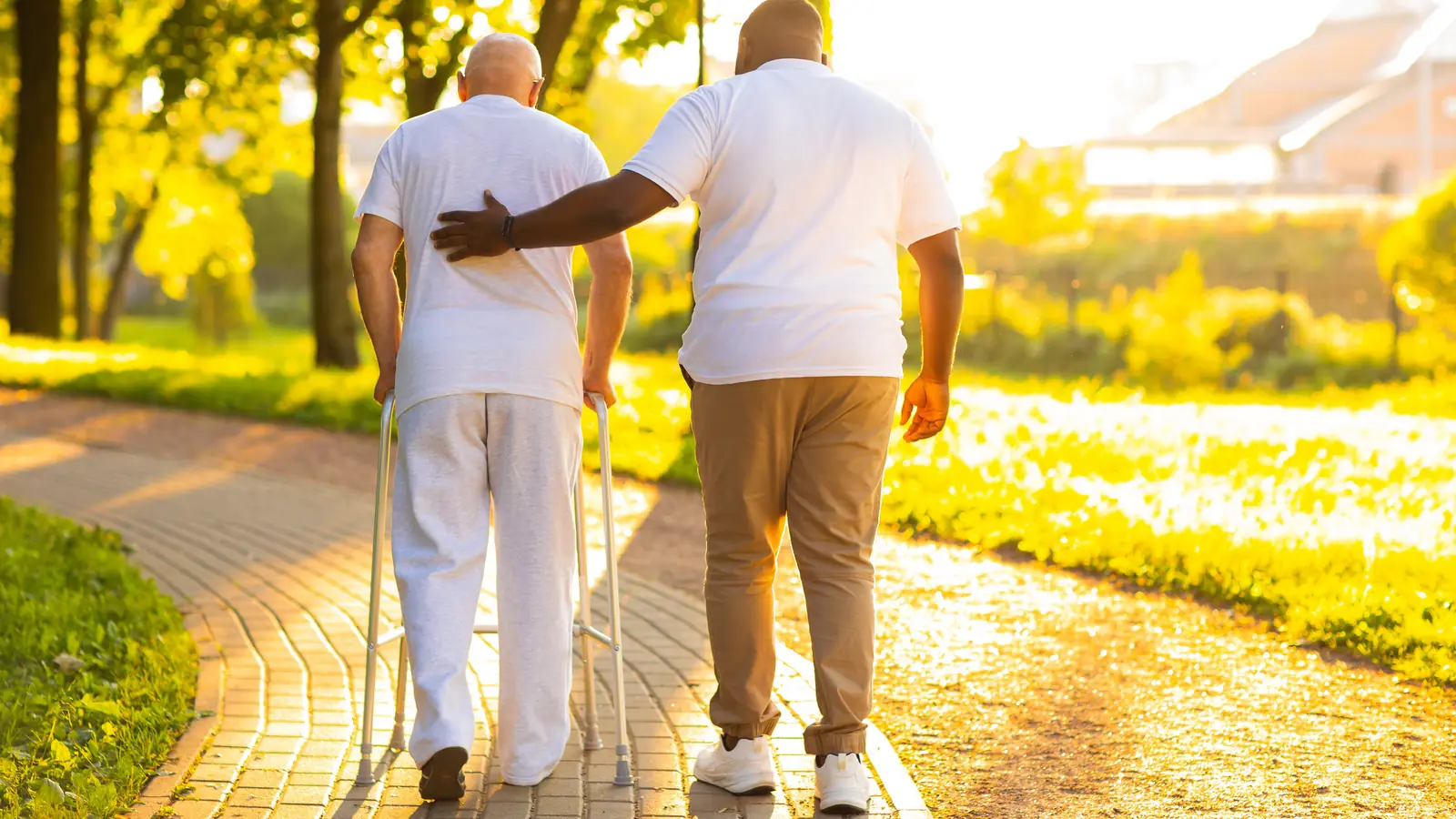 Caregiver supporting a senior man with a walker on a sunny park path